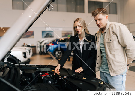 Pensive male customer in casual clothes standing thinking about buying car. Professional female salesperson with client in car showroom near open hood. Confident saleswoman explaining all car features Pensive male customer in casual clothes standing thinking about buying car. Professional female salesperson with client in car showroom near open hood. Confident saleswoman explaining all car features 118952545