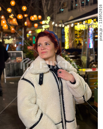 Millennial woman standing outdoors at Christmas night, wrapped in a cozy shearling jacket. the city lights in the background create a warm and Christmas holidays atmosphere, enhancing the peaceful and 118952916