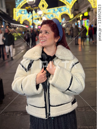 Millennial woman standing outdoors at Christmas night, wrapped in a cozy shearling jacket. the city lights in the background create a warm and Christmas holidays atmosphere, enhancing the peaceful and Millennial woman standing outdoors at Christmas night, wrapped in a cozy shearling jacket. the city lights in the background create a warm and Christmas holidays atmosphere, enhancing the peaceful and 118952923