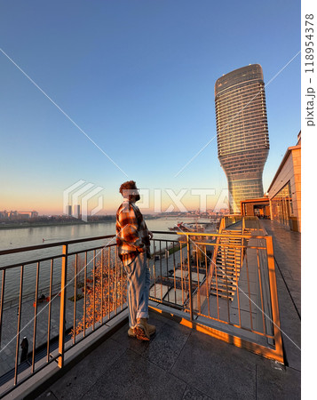 Man stands thoughtfully on a balcony, overlooking a river with a modern skyscraper in Belgrade city during a serene sunset. the warm glow of the evening light casts a peaceful ambiance over the urban Man stands thoughtfully on a balcony, overlooking a river with a modern skyscraper in Belgrade city during a serene sunset. the warm glow of the evening light casts a peaceful ambiance over the urban 118954378