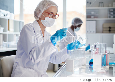 Medium shot of female professional chemist in medical mask dripping liquid from pipette into test tube while conducting chemical experiment in scientific laboratory setting, copy space 118954720