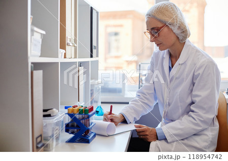 Side view of concentrated female chemist wearing lab coat and hairnet making records of blood test results in notepad while sitting at working table in laboratory, copy space 118954742
