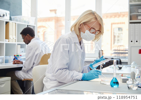 SIde view of female biochemist in protective mask studying bacteria in petri dish working with pipette while sitting at table in laboratory of scientific research service, copy space SIde view of female biochemist in protective mask studying bacteria in petri dish working with pipette while sitting at table in laboratory of scientific research service, copy space 118954751