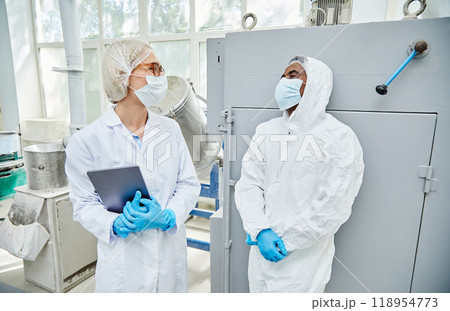 Diverse team of two professional pharmaceutical industry workers in uniform smiling while communicating on production floor at manufacturing plant 118954773