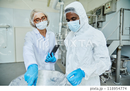 Medium shot of female process supervisor in lab coat pointing at equipment controlling production quality with African American male manufacturing technician, working in protective suit at factory 118954774