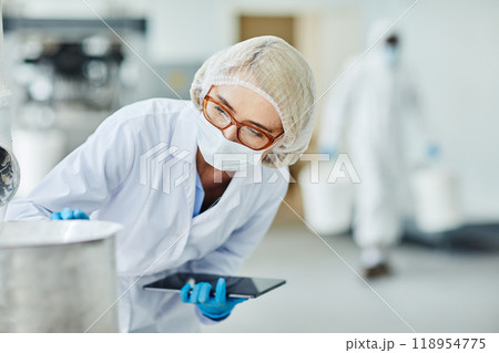 Female manufacturing supervisor in protective mask and glasses overseeing equipment while controlling production process and documenting results in digital tablet at pharmaceutical plant, copy space Female manufacturing supervisor in protective mask and glasses overseeing equipment while controlling production process and documenting results in digital tablet at pharmaceutical plant, copy space 118954775