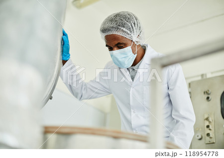 Medium shot of African American male technician in protective mask opening lid of huge steel tank inspecting production process on shop floor of pharmaceutical manufacturing plant, copy space Medium shot of African American male technician in protective mask opening lid of huge steel tank inspecting production process on shop floor of pharmaceutical manufacturing plant, copy space 118954778