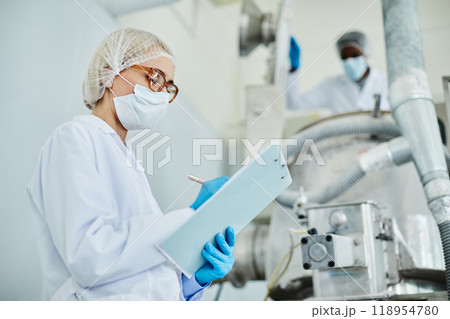 Side view of female technician in protective mask taking notes on notepad while inspecting manufacturing machine on shop floor of pharmaceutical factory, copy space Side view of female technician in protective mask taking notes on notepad while inspecting manufacturing machine on shop floor of pharmaceutical factory, copy space 118954780