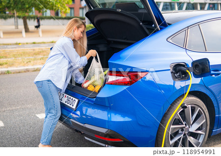 Electric car EV charging while woman put bag with vegetables to the trunk.  118954991