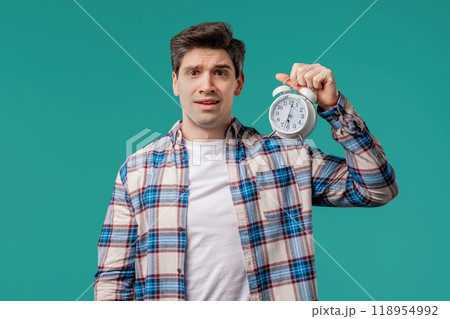 Woken up by alarm clock sleepy young man holding it in hand. Blue background Woken up by alarm clock sleepy young man holding it in hand. Blue background 118954992