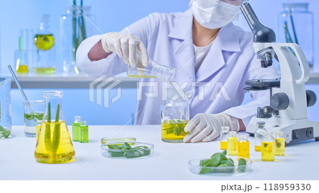 An illustration for a lab template depicts a scientist adding a yellow liquid to a glass aloe vera container. On a table, aloe vera leaves and a green liquid are surrounding the scientist. An illustration for a lab template depicts a scientist adding a yellow liquid to a glass aloe vera container. On a table, aloe vera leaves and a green liquid are surrounding the scientist. 118959330