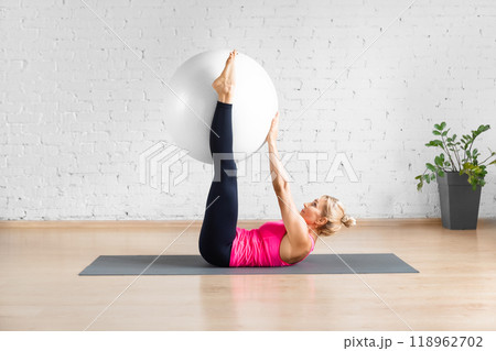 Straight leg hip crunch exercise with fitness ball. Fit Caucasian woman in red tank top does workout with props in loft studio indoor. 118962702