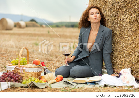 Woman is dressed in jacket with deep neckline, sitting near haystack with glass of wine and book 118962826