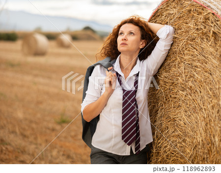 Young woman in white blouse and gray business suit standing on soft plaid mat near stack of straw Young woman in white blouse and gray business suit standing on soft plaid mat near stack of straw 118962893