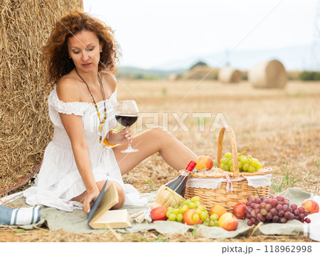 Carefree woman in white sitting on picnic near haystack with wine and book Carefree woman in white sitting on picnic near haystack with wine and book 118962998