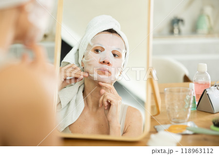 Middle-aged woman applying tissue mask on her facial skin sitting in front of the mirror 118963112