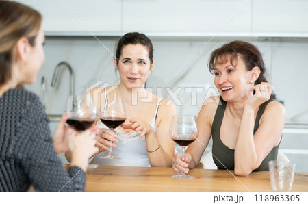 Three happy middle-aged women sitting at table with a glass of wine while having lively conversation Three happy middle-aged women sitting at table with a glass of wine while having lively conversation 118963305