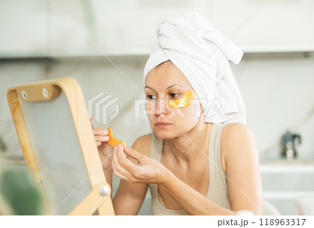 Middle-aged woman applying eye patch mask on her face sitting in front of the mirror Middle-aged woman applying eye patch mask on her face sitting in front of the mirror 118963317