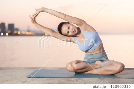 Near sea during sunset, girl performs variations of padmasana with body tilt 118963330