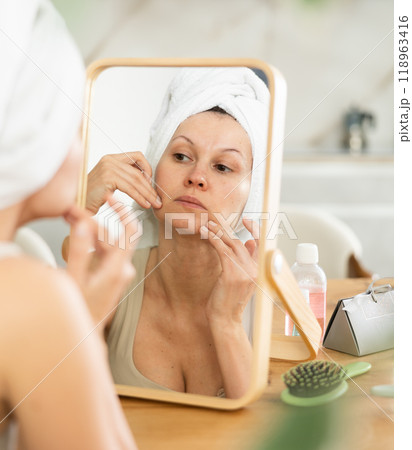 Woman with hair wrapped in towel inspecting complexion in mirror Woman with hair wrapped in towel inspecting complexion in mirror 118963416