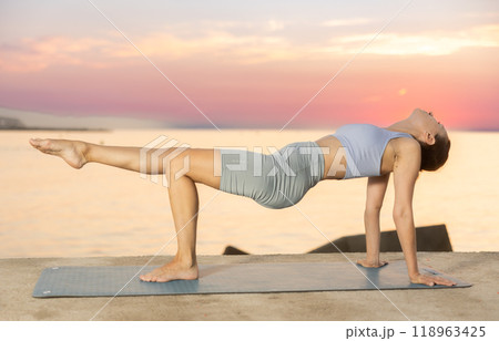 Sporty energetic experienced young woman yoga trainer performing upward plank pose on rug on beach at sunrise. Fitness concept 118963425