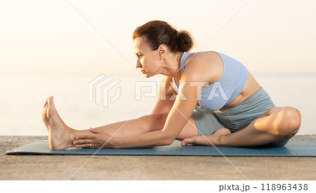 Near sea during sunset, girl performs variations of Janu Shirshasana 118963438