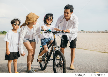 Under golden hues of summer sunset happy father mother and son share road trip on sandy beach. Teaching and learning bicycle riding become cheerful and heartwarming family experience. Under golden hues of summer sunset happy father mother and son share road trip on sandy beach. Teaching and learning bicycle riding become cheerful and heartwarming family experience. 118970933