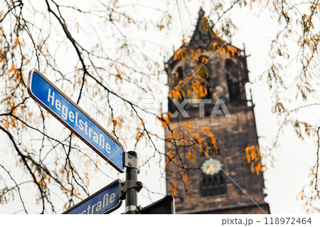 Street name sign Hegelstrasse on blue plate with old historic Magdeburg Cathedral Dom church and autumn trees leaves. Scenic moody street crossing in Germany city. Seasonal weather conditions Street name sign Hegelstrasse on blue plate with old historic Magdeburg Cathedral Dom church and autumn trees leaves. Scenic moody street crossing in Germany city. Seasonal weather conditions 118972464
