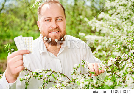 Man allergic suffering from seasonal allergy at spring. Happy man with small white flowers dotted in beard, showing allergy drugs, posing in blossoming garden. Antihistamine medication concept Man allergic suffering from seasonal allergy at spring. Happy man with small white flowers dotted in beard, showing allergy drugs, posing in blossoming garden. Antihistamine medication concept 118973014