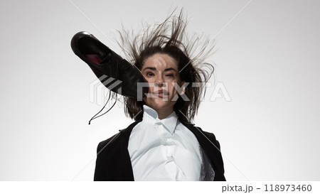 Black leather shoe smashes into woman's cheek against white studio background. Woman in business. Gender roles. 118973460