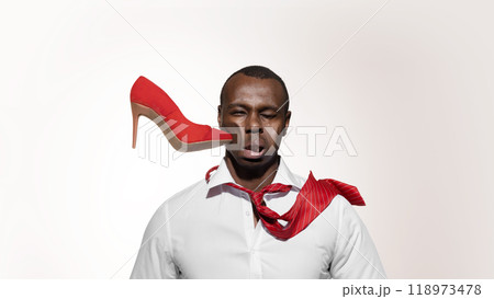 Bright red high heel shoe crashes into African-American man's face, distorting his expression against white studio background. 118973478