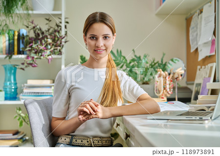 Portrait of smiling female teenager looking at camera, sitting at home at desk with computer Portrait of smiling female teenager looking at camera, sitting at home at desk with computer 118973891