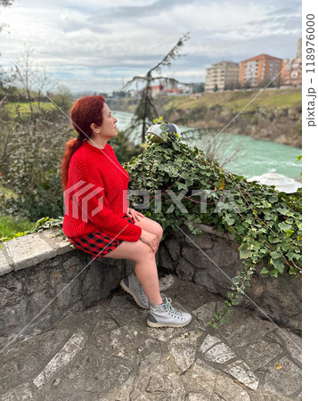 Millennial woman in a red sweater sitting on a stone ledge, enjoying a tranquil river view with a backdrop of city buildings. Autumn scene captures the essence of urban nature and peaceful Millennial woman in a red sweater sitting on a stone ledge, enjoying a tranquil river view with a backdrop of city buildings. Autumn scene captures the essence of urban nature and peaceful 118976000