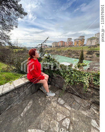 Millennial woman in a red sweater sitting on a stone ledge, enjoying a tranquil river view with a backdrop of city buildings. Autumn scene captures the essence of urban nature and peaceful 118976001
