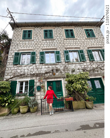 Charming stone building with green shutters and millennial woman in red standing in front. a picturesque scene capturing the essence of rustic architecture and fashion. Travel and journey concept 118976621