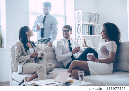 Coffee break. Group of four young people holding coffee cups and discussing something with smile while sitting on the couch at office Coffee break. Group of four young people holding coffee cups and discussing something with smile while sitting on the couch at office 118977525