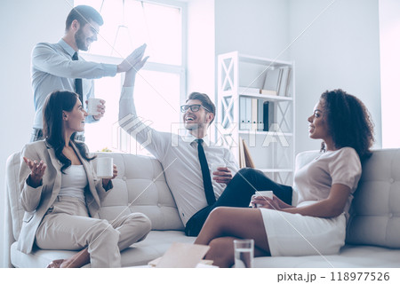 They deserve a little rest. Two handsome man giving high-five and holding coffee cups with smile while sitting on the couch at office with their beautiful coworkers 118977526