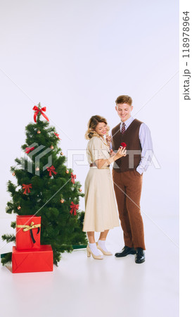 Woman shows man small red gift box, standing by Christmas tree, surrounded by holiday cheer, as they share loving moment. Woman shows man small red gift box, standing by Christmas tree, surrounded by holiday cheer, as they share loving moment. 118978964