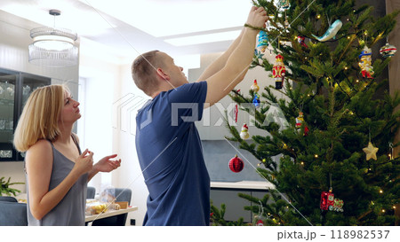 A couple decorates a Christmas tree at home, sharing joy and love in a cozy festive atmosphere. New Year's Eve with family, newlyweds decorate a festive tree A couple decorates a Christmas tree at home, sharing joy and love in a cozy festive atmosphere. New Year's Eve with family, newlyweds decorate a festive tree 118982537