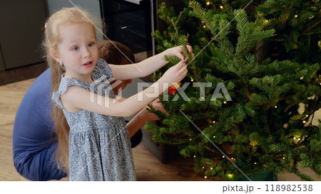 Young girl delicately hanging a christmas ornament on a tree branch with her father's assistance. Little girl decorating Christmas tree with vintage toys Young girl delicately hanging a christmas ornament on a tree branch with her father's assistance. Little girl decorating Christmas tree with vintage toys 118982538