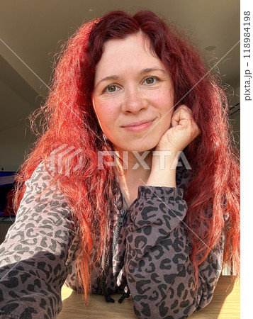 Portrait smiling woman with vibrant red hair basks in the glow of sunlight. her relaxed expression and comfortable attire create an atmosphere of warmth and contentment. 118984198