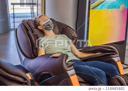 Beautiful young woman wearing a medical mask due to the coronavirus COVID 19 relaxing on the massage chair in airport or in the mall 118985882