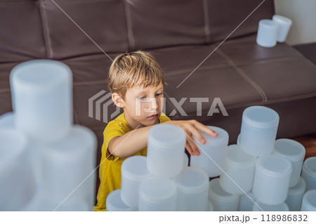 Boy playing with cans of yogurt surrounded by many jars of yoghurt that he uses in a year. A huge amount of plastic food jars. Excessive consumption of plastic. Reduce, reuse, recycle 118986832