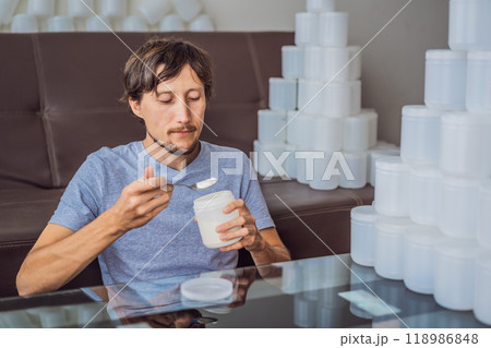 The man eats yogurt and is surrounded by many jars of yoghurt that he uses in a year. A huge amount of plastic food jars. Excessive consumption of plastic. Reduce, reuse, recycle 118986848