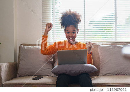 African American woman excitedly celebrates success while sitting on couch with laptop in her home. African American woman excitedly celebrates success while sitting on couch with laptop in her home. 118987061