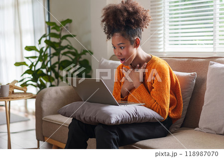 Excited African American young woman engaged in online activity at home in a cozy living room. Excited African American young woman engaged in online activity at home in a cozy living room. 118987070