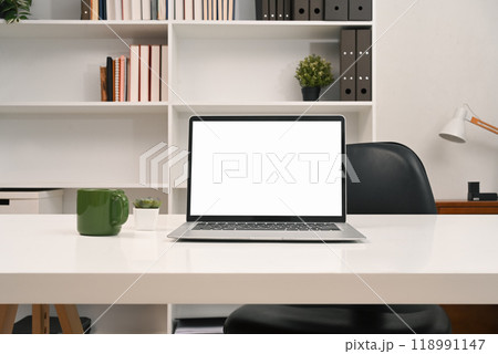 A laptop computer with empty display and office supplies on a desk in front of a bookshelf A laptop computer with empty display and office supplies on a desk in front of a bookshelf 118991147
