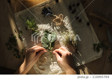 Top view of human hands tying twine around sage sprigs. Linen tablecloth on a wooden table with various herbs. Autumn atmosphere 118995769