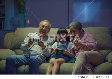Happy Asian family bonding over a video game on a cozy couch, little girl playing video games console with senior grandparents together with controller joysticks while sitting on the sofa at night Happy Asian family bonding over a video game on a cozy couch, little girl playing video games console with senior grandparents together with controller joysticks while sitting on the sofa at night 118998241