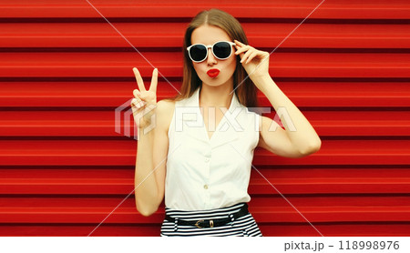 Portrait of stylish young woman model posing blowing kiss wearing sunglasses on red background 118998976